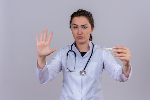 Looking at camera young doctor wearing medical gown wearing stethoscope holding thermometer and shows stop gesture on white wall
