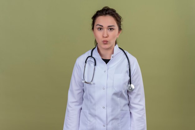 Looking at camera young doctor wearing medical gown wearing stethoscope showing kiss gesture on green wall