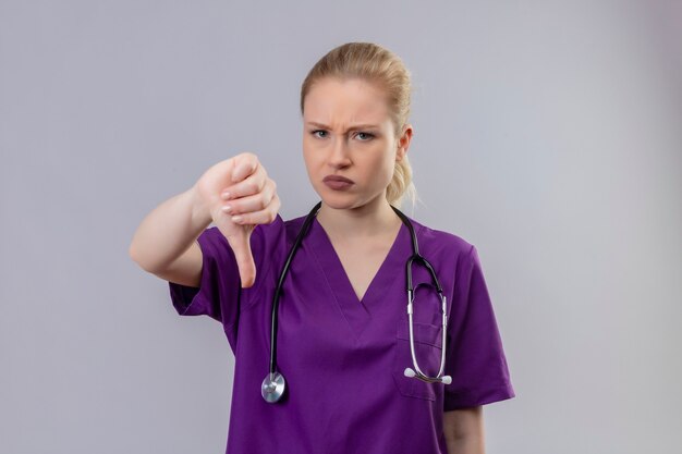 紫色の医療用ガウンと聴診器を身に着けているカメラの若い医者を見て、孤立した白い壁に親指を下ろします