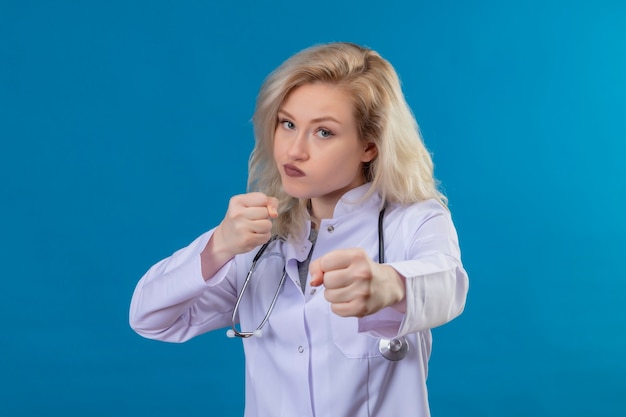 Looking at camera young doctor wearing stethoscope in medical gown standing in fughting pose on blue wall