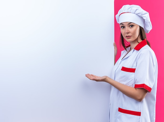 Looking at camera young female cook wearing chef uniform stands nearby white wall and points at wall with hand isolated on pink background with copy space