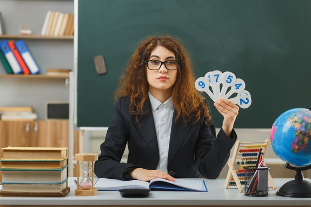 教室で学校の道具を持って机に座っている数のファンを保持している眼鏡をかけている若い女教師のカメラを見て