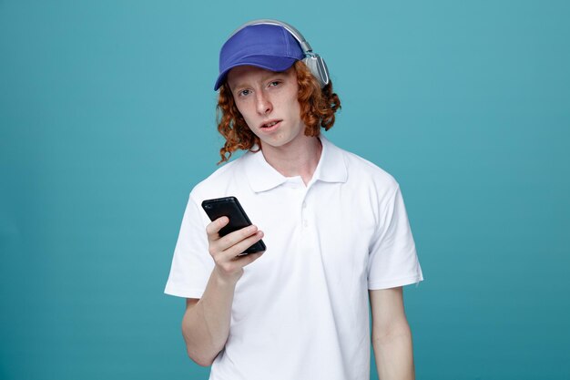 Looking at camera young handsome guy in cap wearing headphones holding phone isolated on blue background
