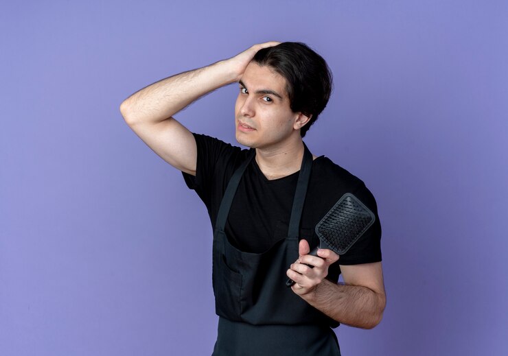 मुफ़्त फोटो looking at camera young handsome male barber in uniform holding comb and putting hand on head isolated on blue background