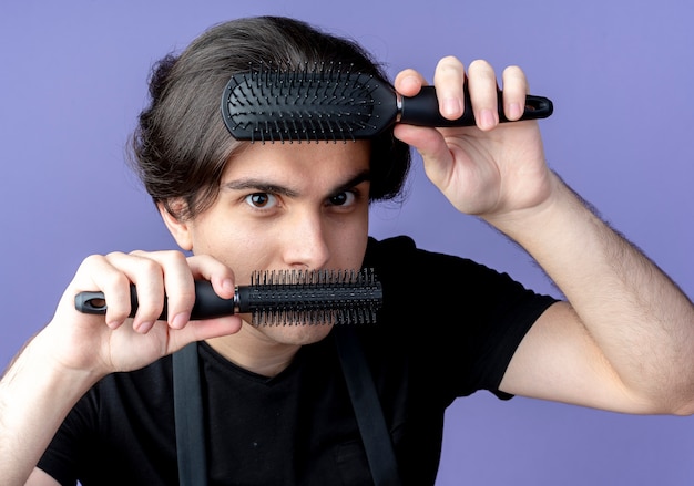 Foto gratuita guardando la fotocamera giovane bel maschio barbiere in uniforme che tiene pettini sulla fronte e sulla bocca isolato su sfondo blu