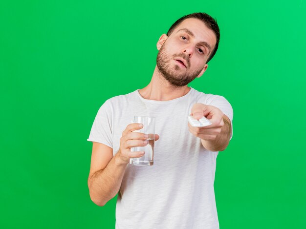 Looking at camera young ill man holding glass of water and holding out pills at camera isolated on green background