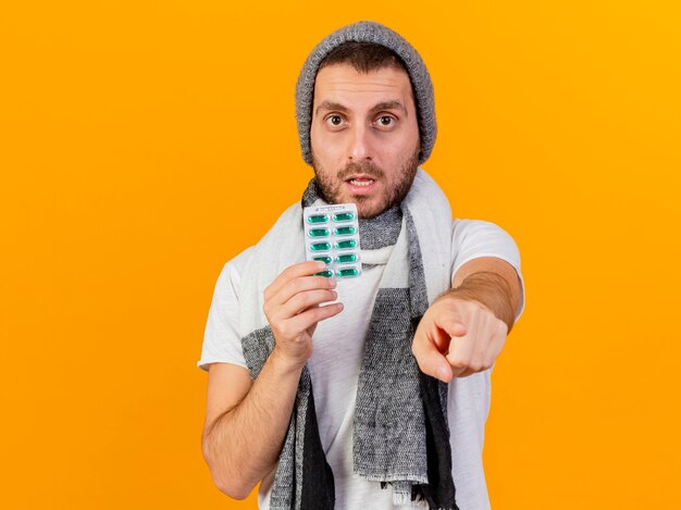 Looking at camera young ill man wearing winter hat and scarf holding pills and showing you gesture isolated on yellow