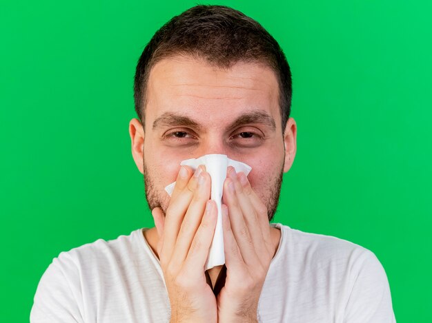 Looking at camera young ill man wiping nose with napkin isolated on green background