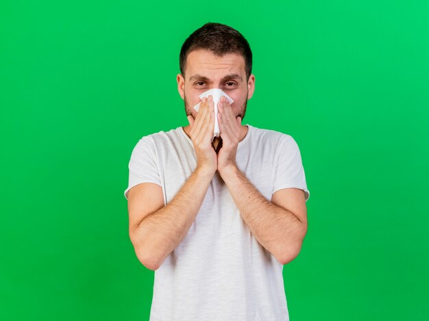 Looking at camera young ill man wiping nose with napkin isolated on green background