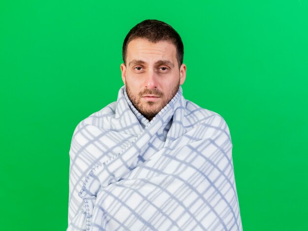 Looking at camera young ill man wrapped in plaid isolated on green background
