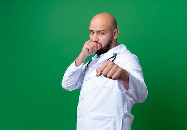 Looking at camera young male doctor wearing medical robe and stethoscope standing in fighting pose isolated on green background with copy space