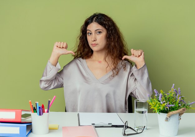 カメラを見て、オフィスツールで机に座っている若いきれいな女性のサラリーマンは、オリーブ色の背景に分離された自分を指しています