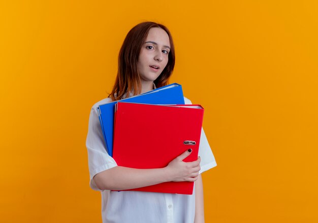 Looking at camera young redhead girl holding folders isolated on yellow background