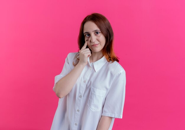 Looking at camera young redhead girl putting finger on eye isolated on pink background