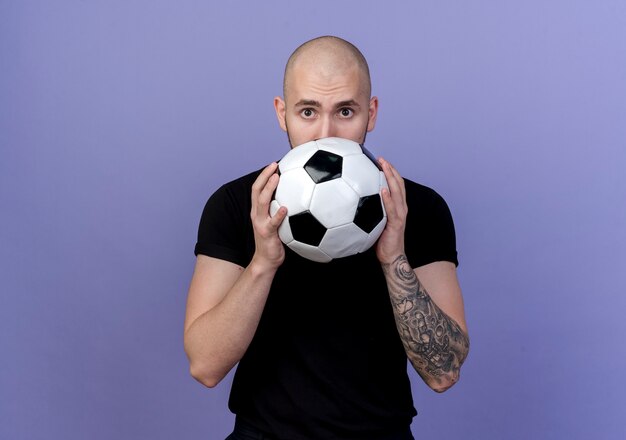 Looking at camera young sporty man covered face with ball isolated on purple background