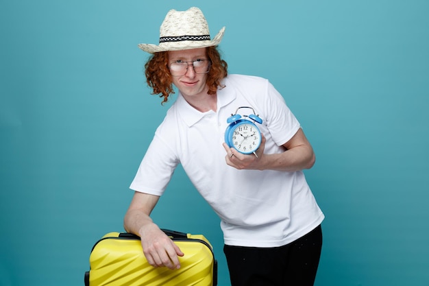 Guardando la fotocamera giovane uomo in viaggio che indossa il cappello che tiene la valigia con sveglia isolata su sfondo blu