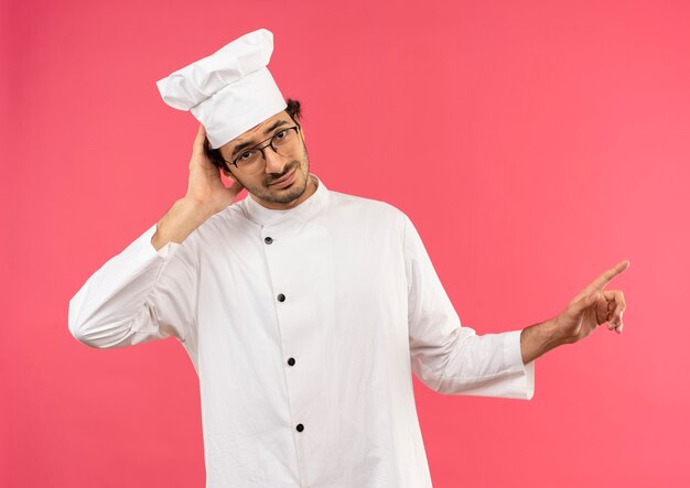 シェフの制服を着た若い男性料理人が混乱し、眼鏡をかけて頭に手を当て、横を指している