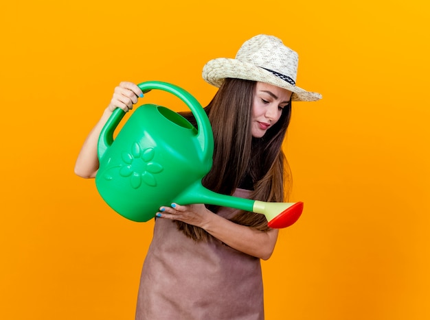 Looking at down beautiful gardener girl wearing uniform and gardening hat watering with watering can isolated on orange background