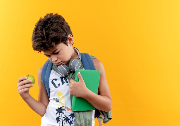 コピースペースで黄色の背景に分離されたリンゴと本を保持しているバックバッグとヘッドフォンを身に着けている悲しい小さな男子生徒を見下ろします