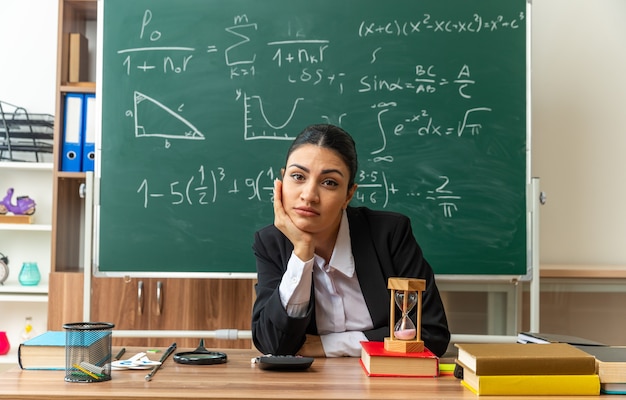 Free photo looking at front young female teacher sits at table with school supplies putting hand on chin in classroom