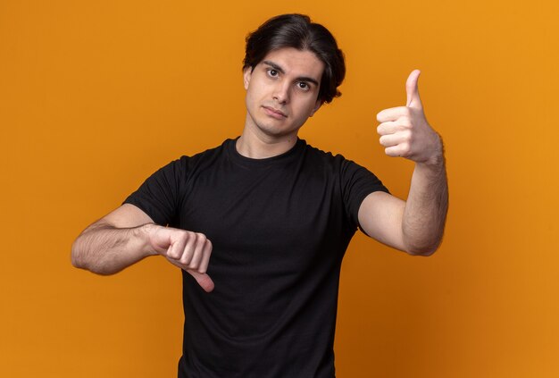 Looking at front young handsome guy wearing black t-shirt showing thumbs up and down isolated on orange wall