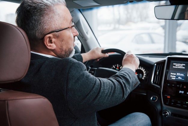 フロントミラーをのぞき込む。現代の新しい車を運転する公式の服の上級ビジネスマンの後ろからの眺め