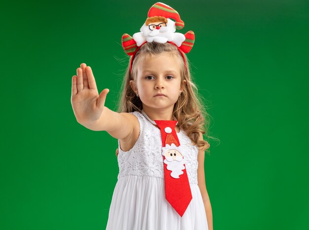 Looking  little girl wearing christmas hair hoop with tie showing stop gesture isolated on green wall