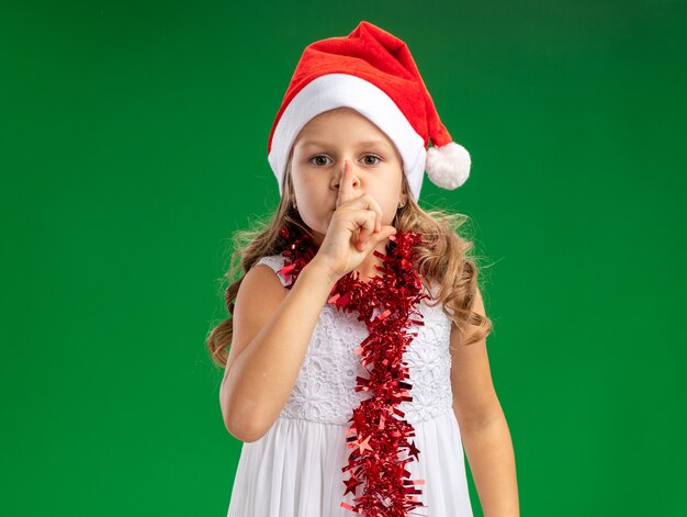 Looking  little girl wearing christmas hat with garland on neck showing silence gesture isolated on green wall