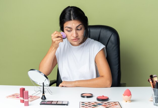 Looking at mirror beautiful woman sits at table with makeup tools applying tone cream with sponge 