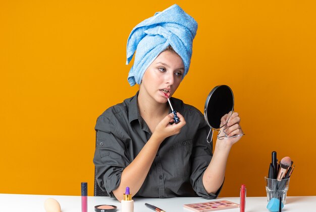 Foto gratuita guardando allo specchio la bella donna si siede al tavolo con gli strumenti per il trucco avvolti i capelli in un asciugamano applicando lucidalabbra