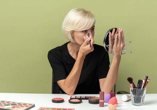 Looking at mirror young beautiful girl sits at table with makeup tools applying eyeshadow isolated on olive green wall