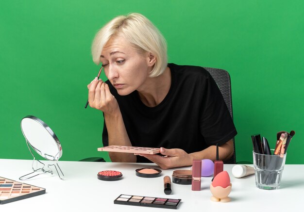 Looking at mirror young beautiful girl sits at table with makeup tools applying eyeshadow with makeup brush isolated on green wall