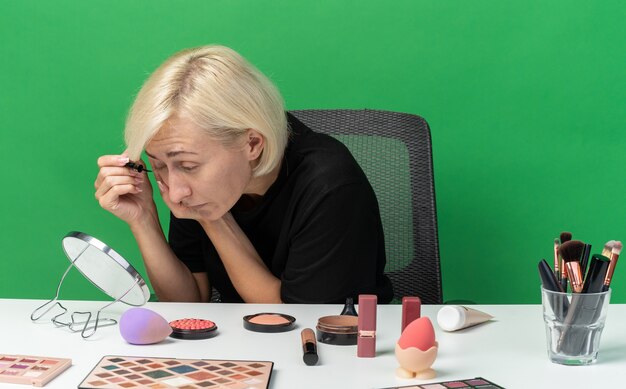 Looking at mirror young beautiful girl sits at table with makeup tools applying mascara isolated on green wall