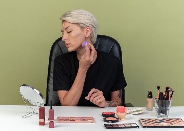 Looking at mirror young beautiful girl sits at table with makeup tools applying tone-up cream with sponge isolated on olive green wall