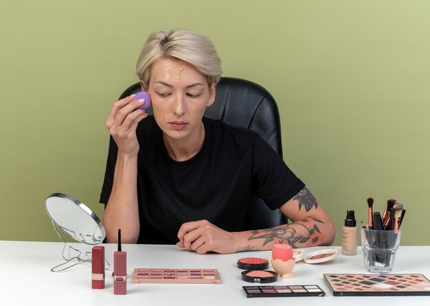 Looking at mirror young beautiful girl sits at table with makeup tools applying tone-up cream with sponge isolated on olive green wall