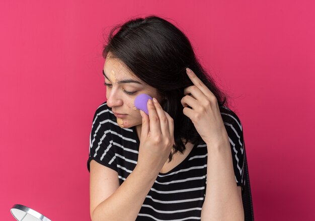 Guardando lo specchio la giovane bella ragazza si siede al tavolo con gli strumenti per il trucco applicando la crema tonificante con la spugna isolata sulla parete rosa pink