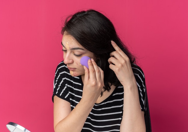 Looking at mirror young beautiful girl sits at table with makeup tools applying tone-up cream with sponge isolated on pink wall