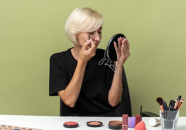 Looking at mirror young beautiful girl sits at table with makeup tools draw arrow with eyeliner isolated on olive green wall