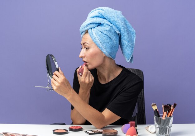 Looking at mirror young beautiful girl sits at table with makeup tools wiping hair in towel applying lipstick isolated on blue wall