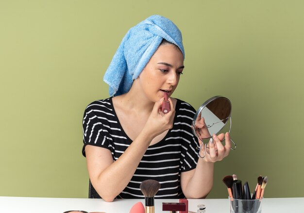 Looking at mirror young beautiful girl sits at table with makeup tools wrapped hair in towel applying lipstick isolated on olive green wall