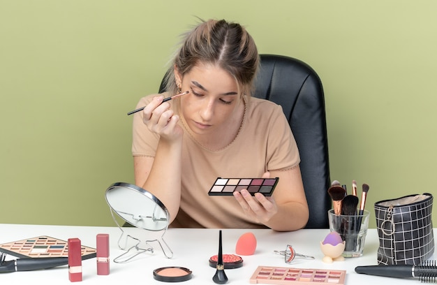 Looking at mirror young beautiful girl sitting at desk with makeup tools applying eyeshadow with makeup brush isolated on olive green wall