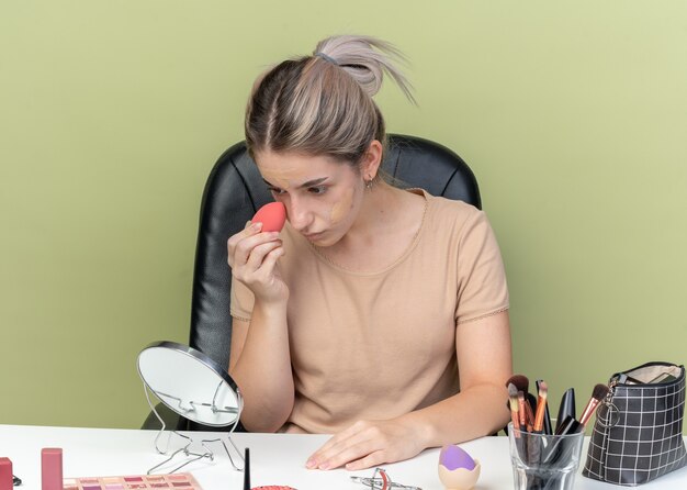 Looking at mirror young beautiful girl sitting at desk with makeup tools wiping tone-up cream with sponge isolated on olive green wall