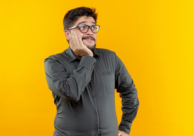 Looking at side amiling young businessman wearing glasses putting hand under chin isolated on yellow background