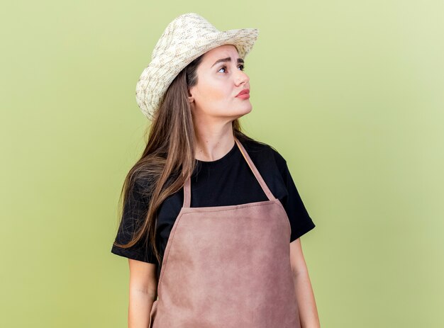 Looking at side beautiful gardener girl in uniform wearing gardening hat isolated on olive green background