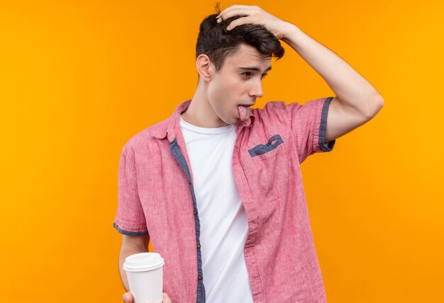 Глядя на бок кавказского молодого человека в розовой рубашке, держащего чашку кофе, показывая язык и положившего руку на голову на изолированной оранжевой стене