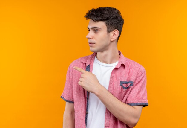 Looking at side caucasian young man wearing pink shirt points to side on isolated orange wall