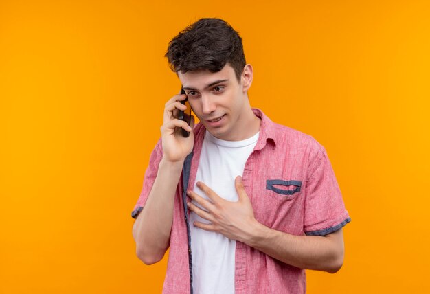 ピンクのシャツを着ている白人の若い男が孤立したオレンジ色の壁に電話で話す側を見て