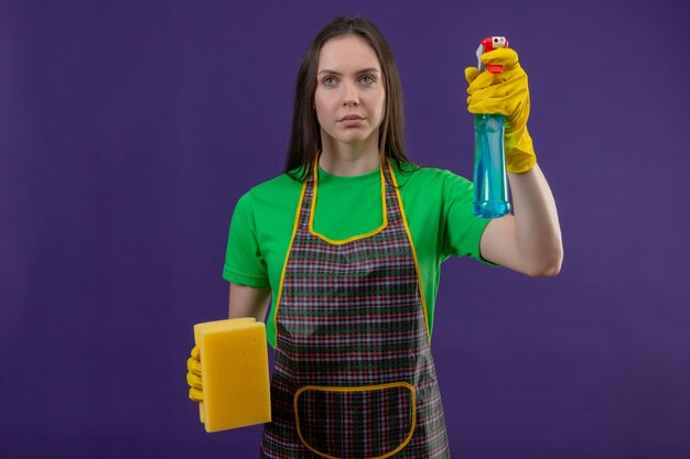 Looking at side cleaning young woman wearing uniform in gloves holding cleaning spray and sponge on purple wall