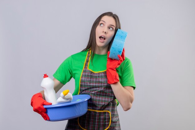 Looking at side cleaning young woman wearing uniform in red gloves holding cleaning tools with sponge on isolated white wall