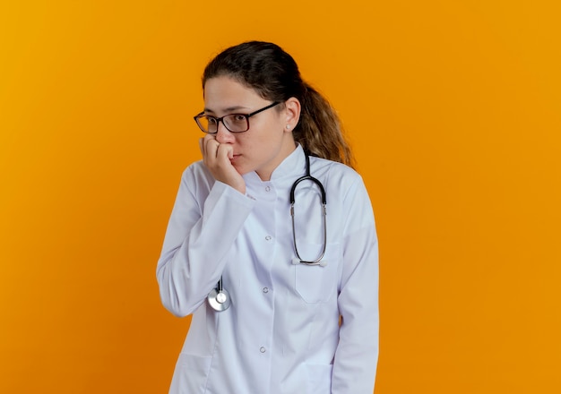 Guardando il lato interessato giovane dottoressa che indossa veste medica e stetoscopio con gli occhiali morde le unghie isolate sulla parete arancione